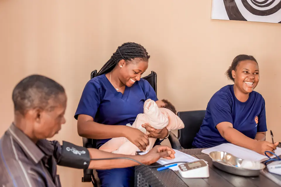 A nurse holds a newborn while a colleague takes an older patient's blood pressure.