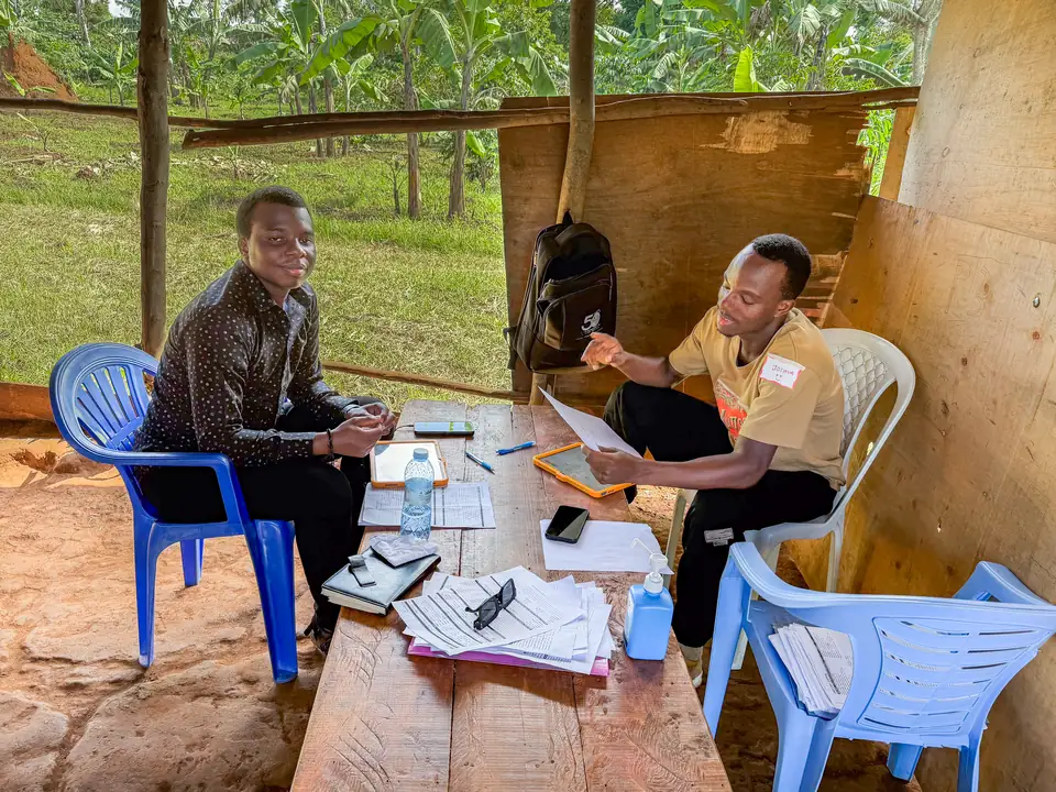 Two clinic staff register patients on tablets at a wooden table in an open-air shelter.