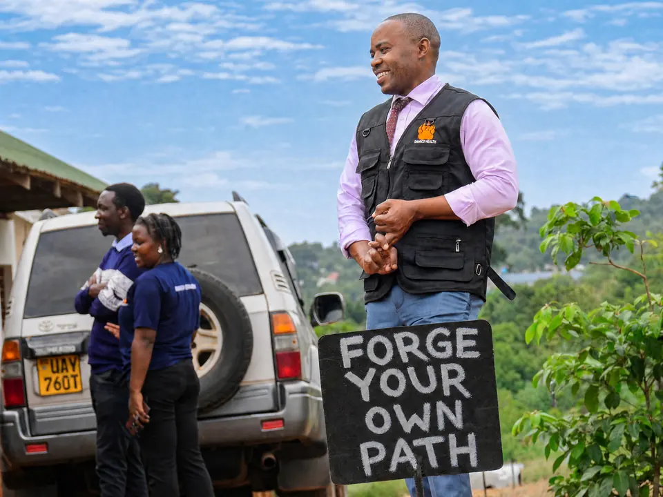 Dr. Kansiime Erhardt, in a Damico Health field vest, stands beside a hand-painted sign that reads Forge your own path.
