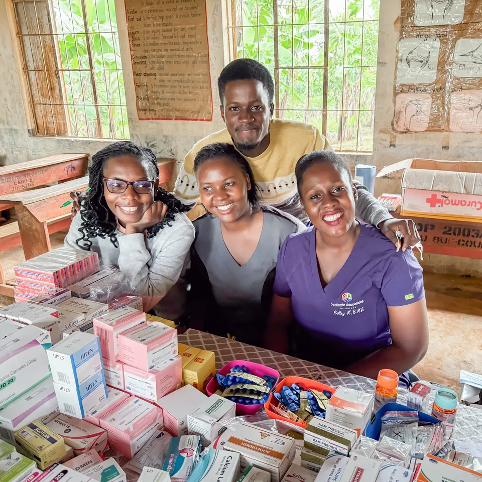 Four clinic staff smile behind a table stacked with boxes of medication in a village classroom.